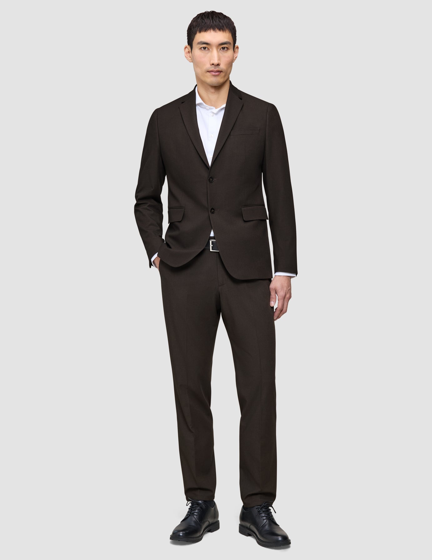 En mand er klædt i The suit that does it all i mørkebrun, en hvid skjorte med høj krave og sorte lædersko.