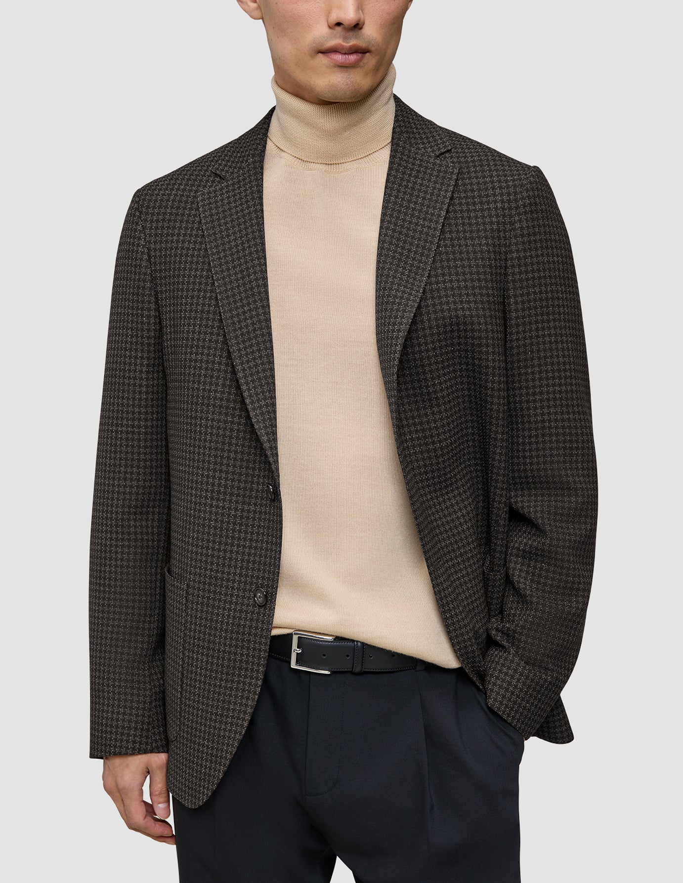 En mand har en mørkebrun, slim-fit Timeless-uldblazer med tern på over en beige rullekravesweater og sorte bukser.
