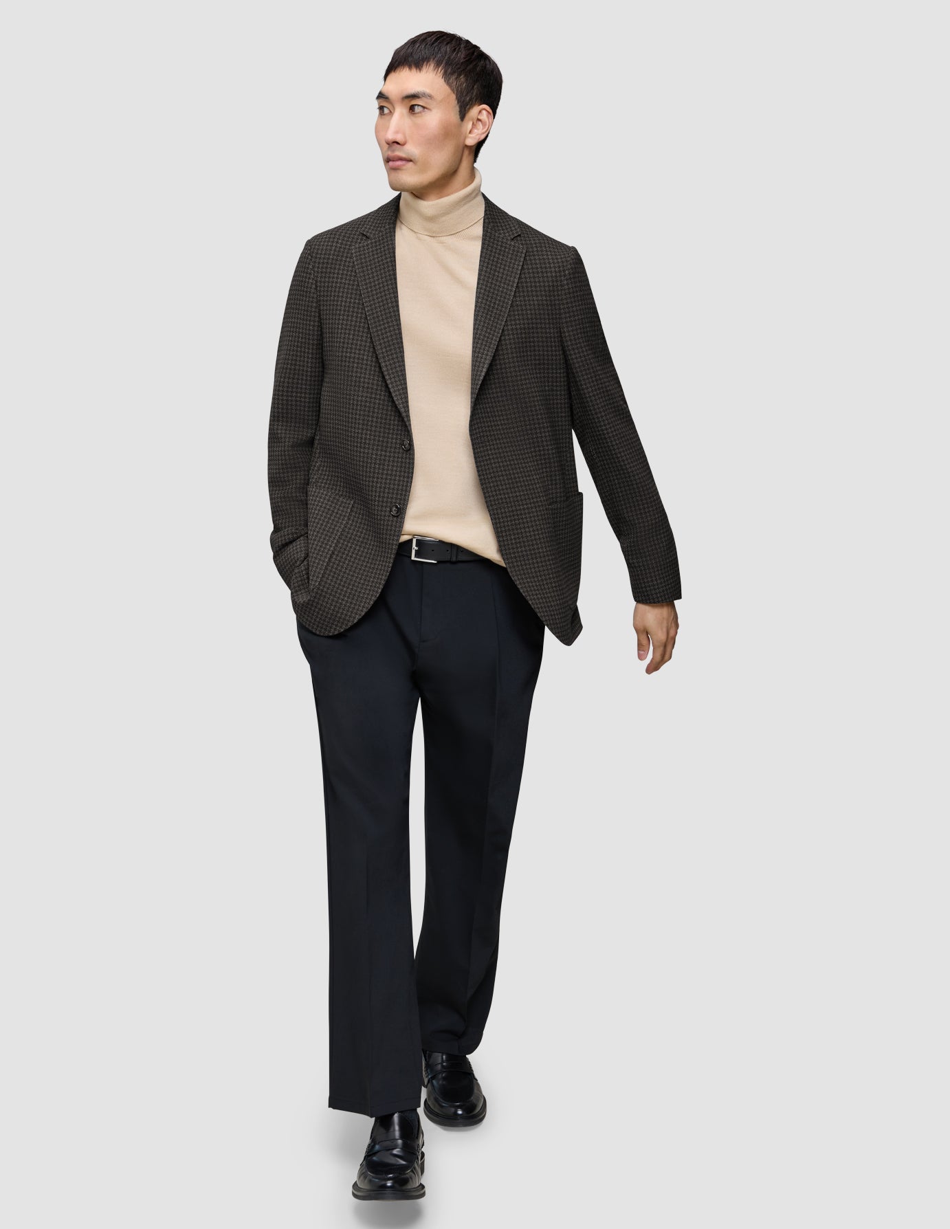 En mand har en mørkebrun, slim-fit Timeless-uldsblazer med tern på over en beige rullekravebluse og mørke bukser.