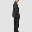 En mand står med siden til i en Timeless-uldsblazer i slim-fit med mørkebrunt ternet mønster, over en beige rullekravebluse og mørke bukser.