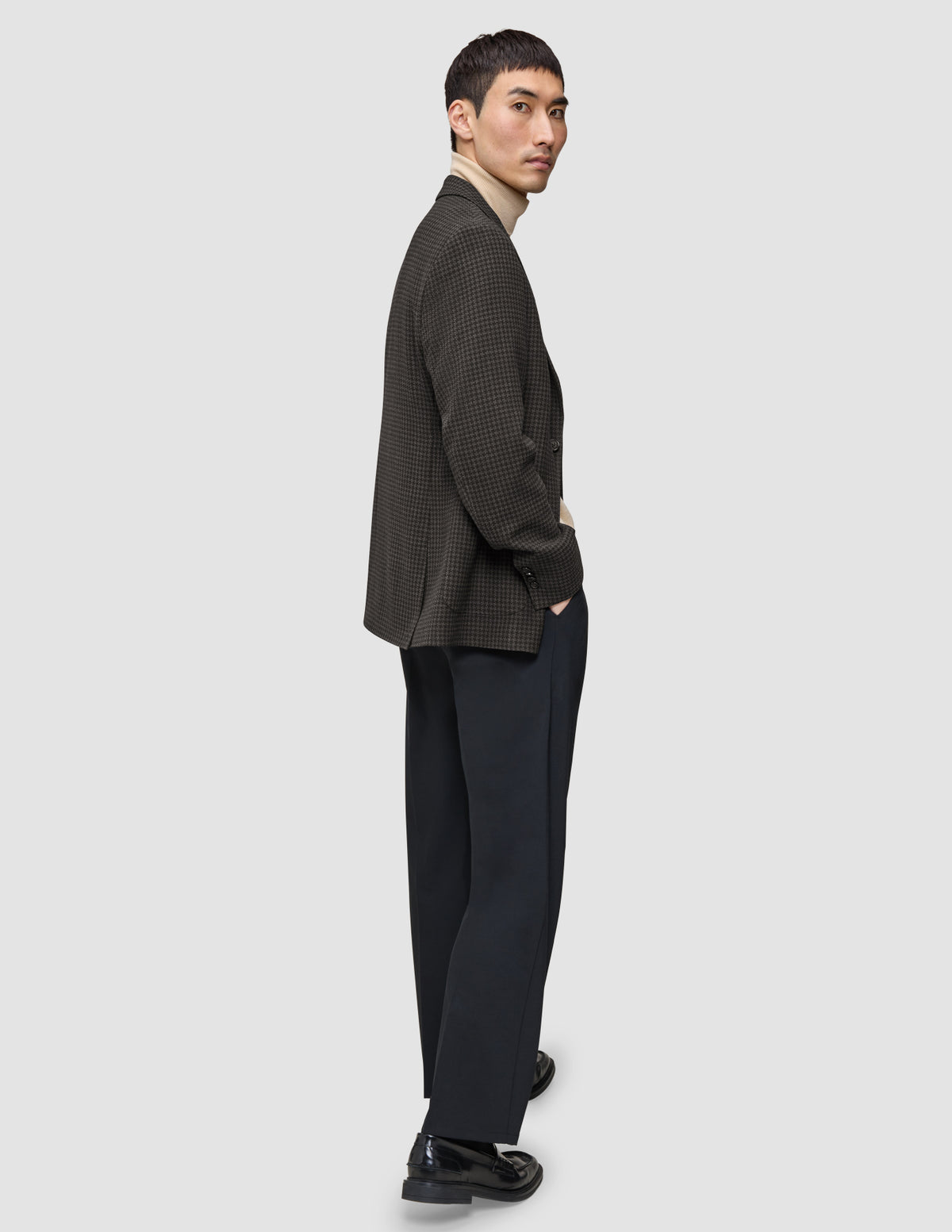 En mand står med siden til i en Timeless-uldsblazer i slim-fit med mørkebrunt ternet mønster, over en beige rullekravebluse og mørke bukser.