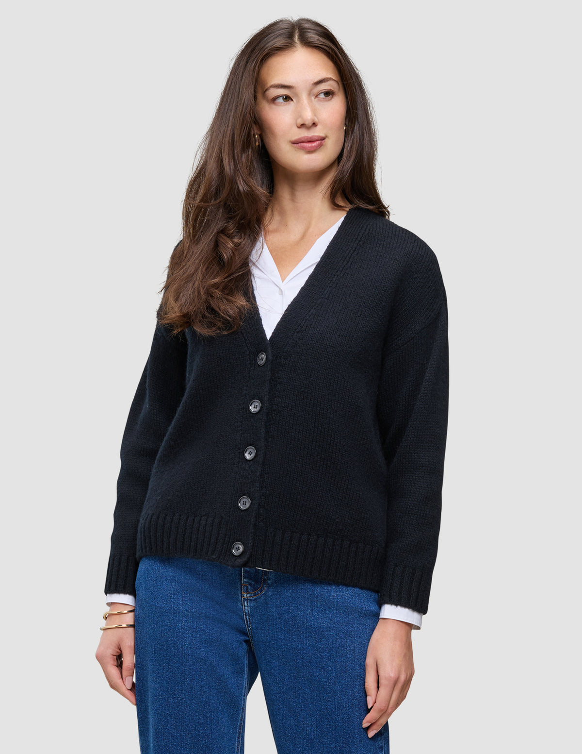 En kvinde iført en sort, tyk, blød uldstrikket cardigan fra serien Women over en hvid skjorte og blå jeans.