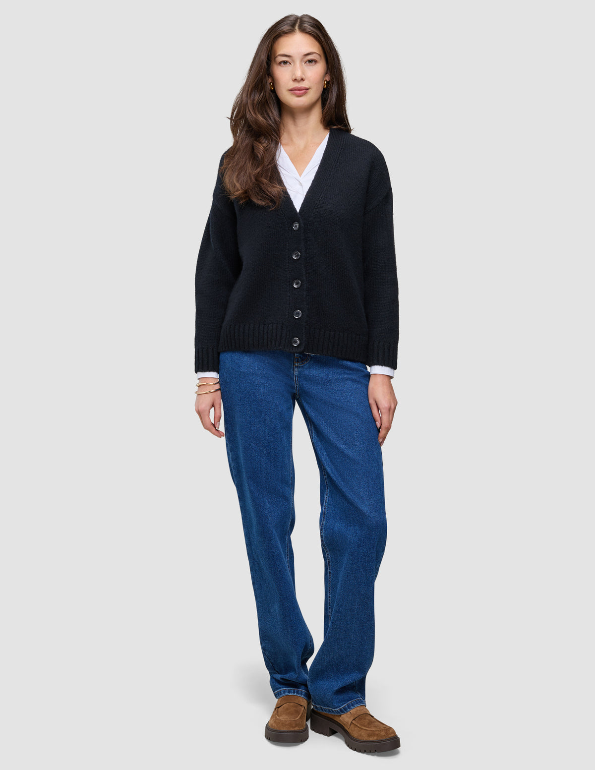 En kvinde iført en sort, tyk, blød uldstrikket cardigan fra serien Women over en hvid skjorte med krave og blå jeans.