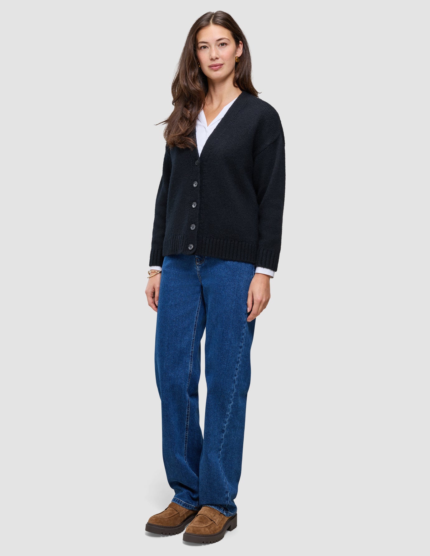 En kvinde iført en sort, tyk, blød uldstrikket cardigan fra serien Women over en hvid skjorte og blå jeans.