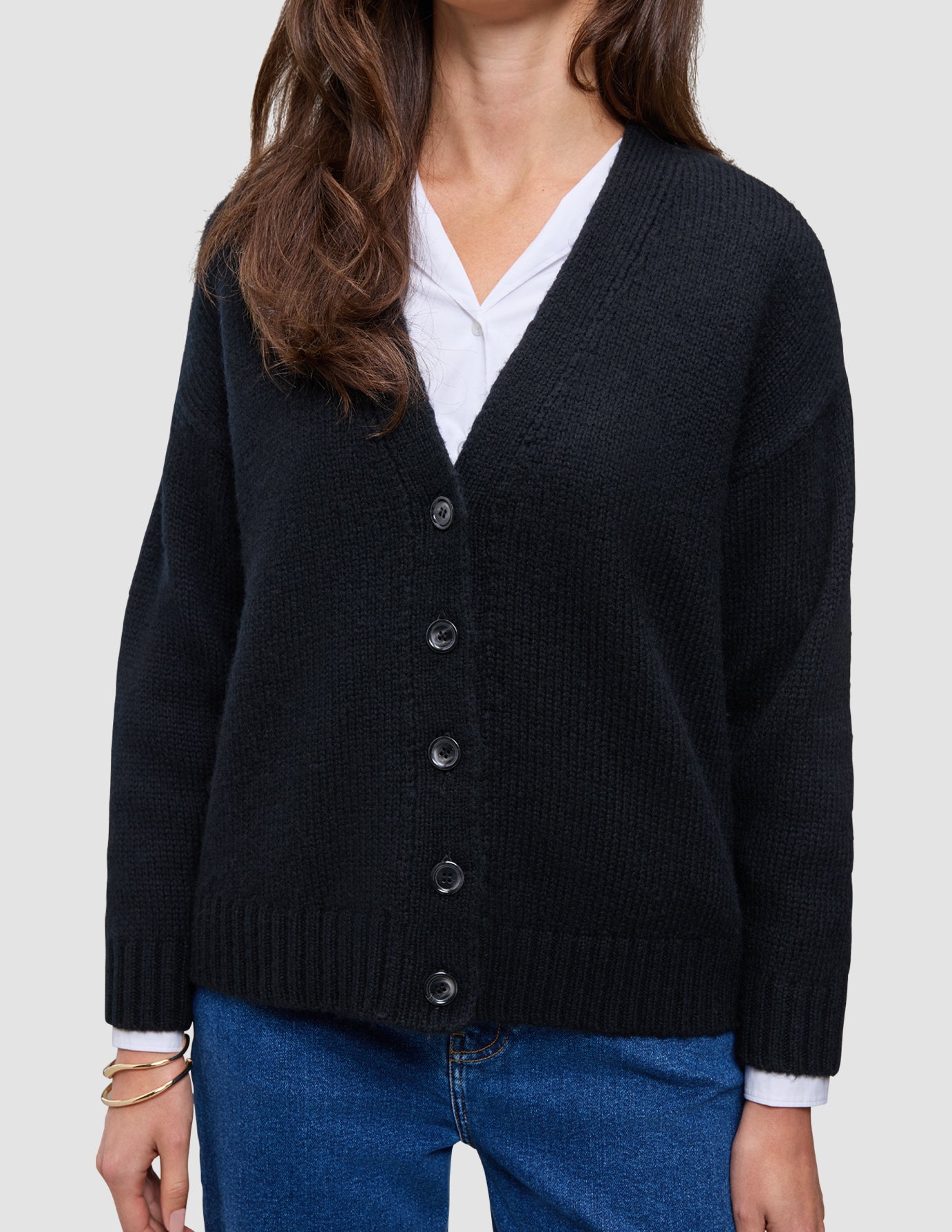 En person, der har en sort, tyk, blød uldstrikket cardigan til kvinder på over en hvid skjorte med krave og blå jeans.