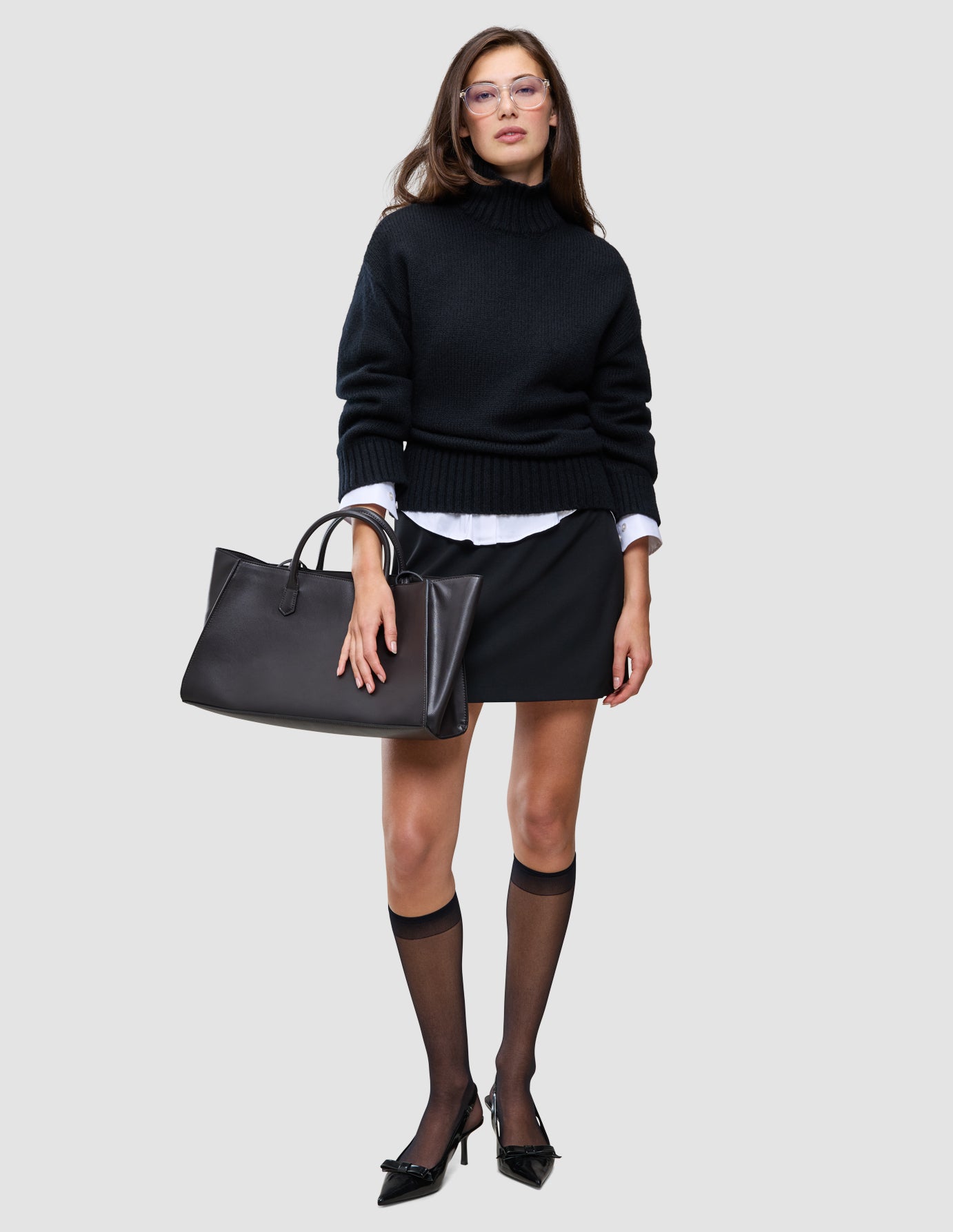 En kvinde har en sort, tyk og blød uldstrikket rullekravebluse til kvinder på over en skjorte med krave, et nederdel, knæstrømper, pumps og en totebag.
