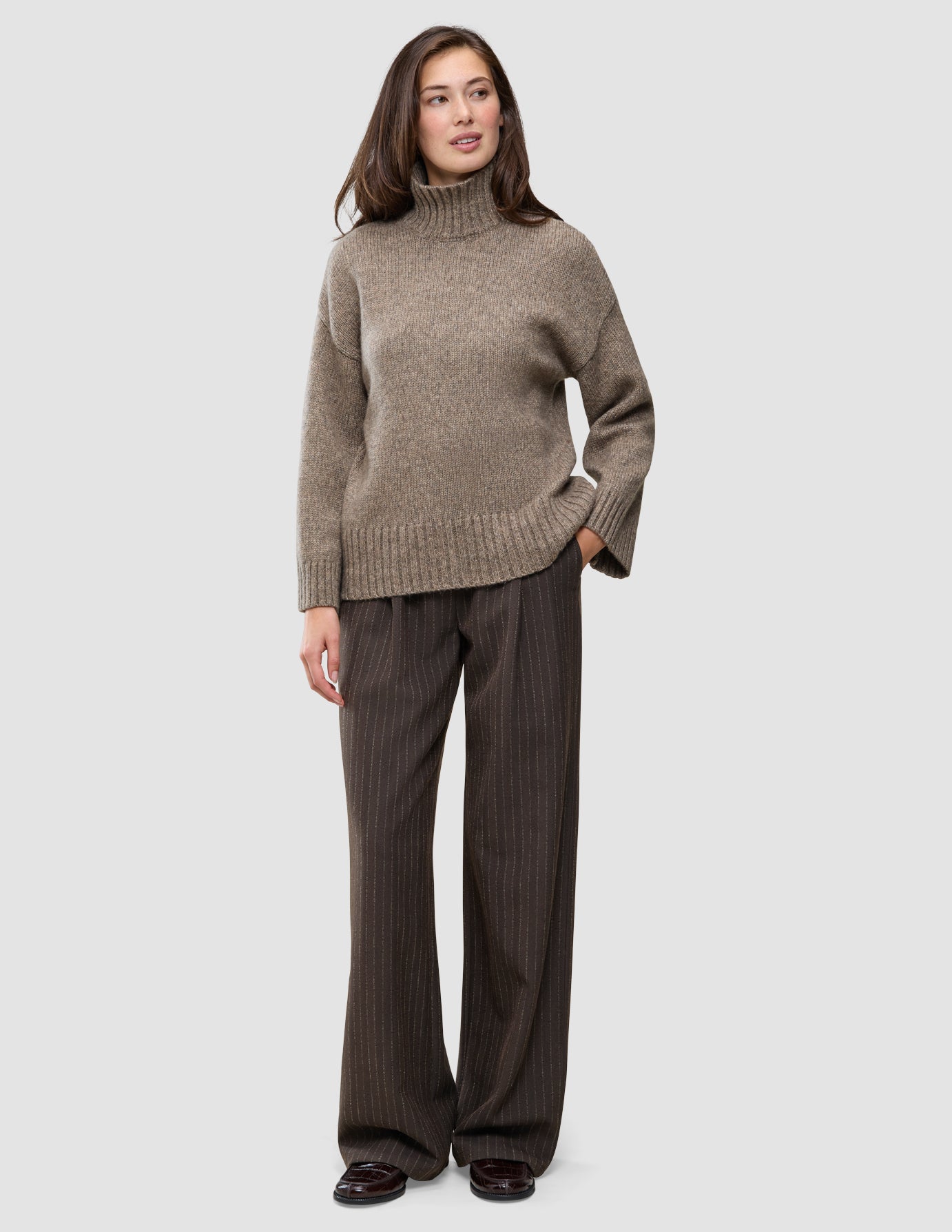 En kvinde iført en Women Chunky Soft Wool Knit Turtleneck Taupe Melange-trøje og mørkebrune bukser med tynde striber.