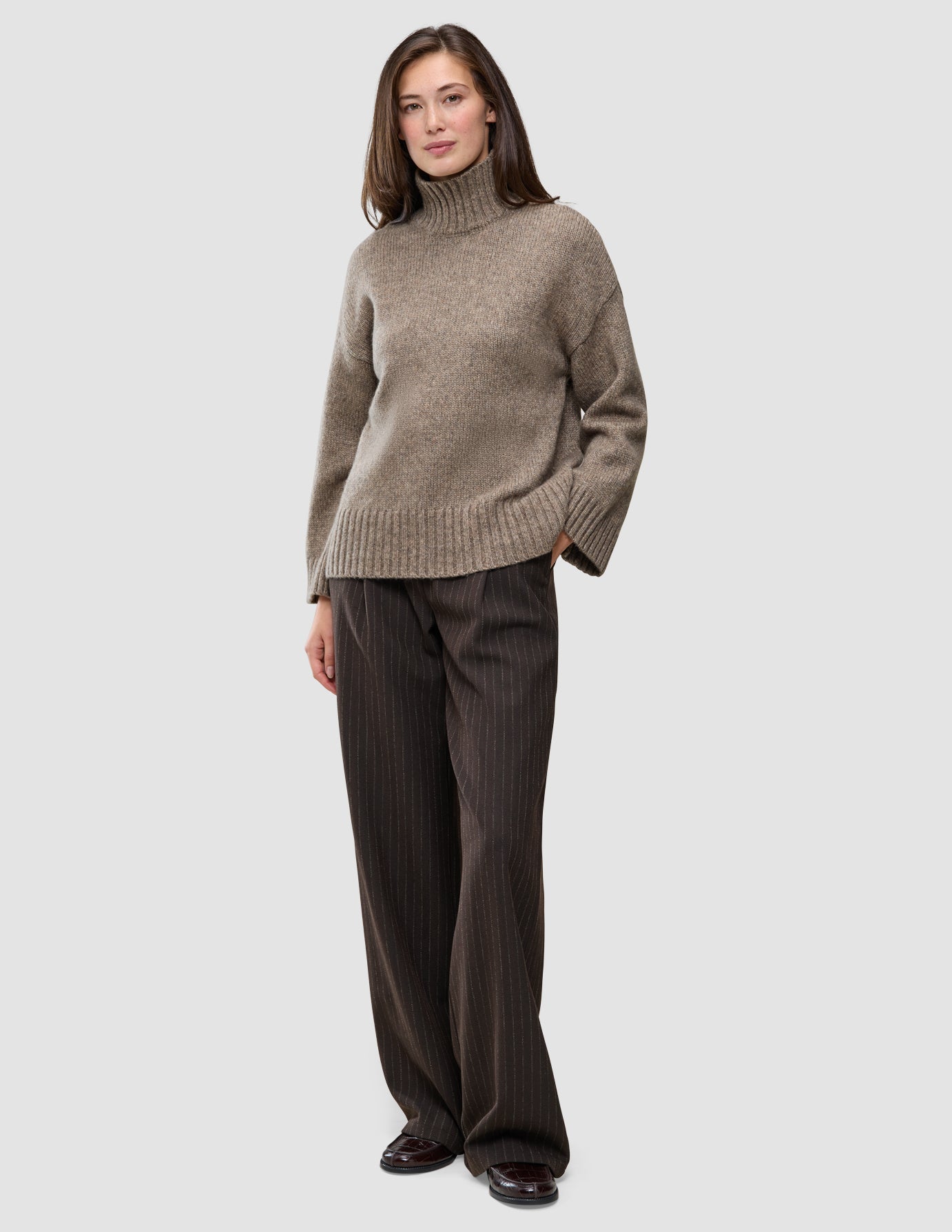 En kvinde iført en Women Chunky Soft Wool Knit Turtleneck Taupe Melange-trøje og mørkebrune bukser med tynde striber.