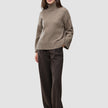 En kvinde iført en Women Chunky Soft Wool Knit Turtleneck Taupe Melange-trøje og mørkebrune bukser med tynde striber.