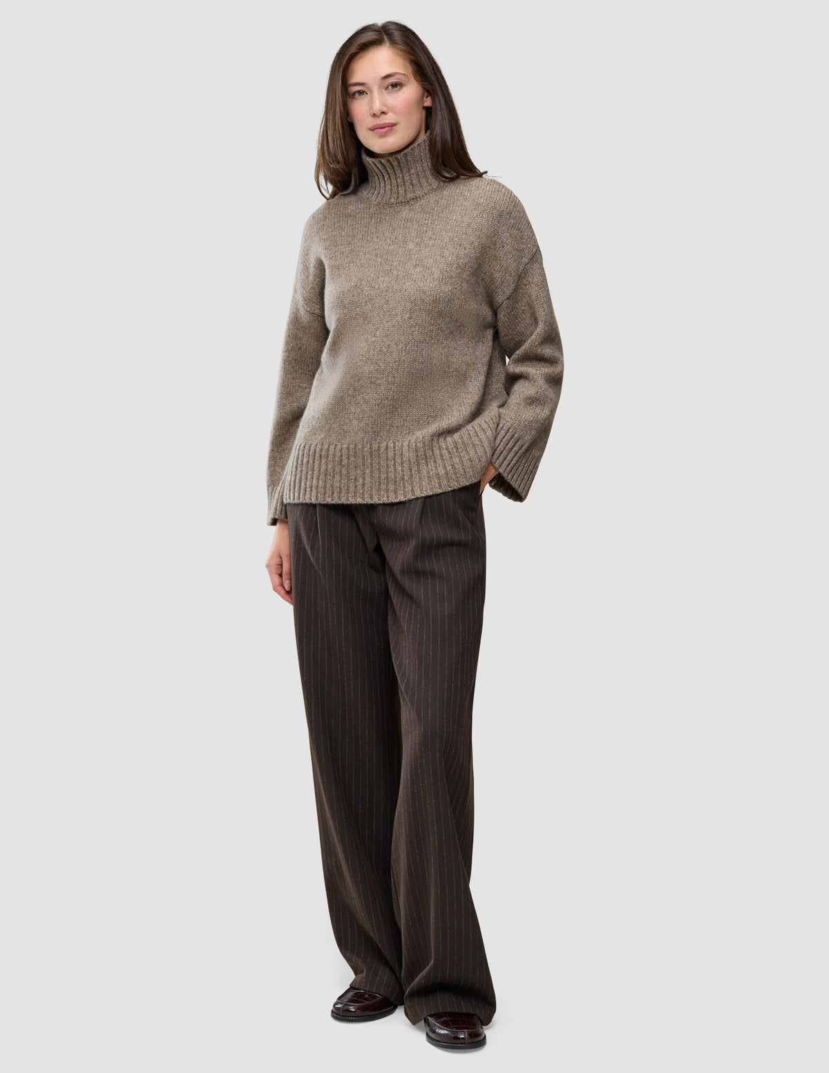 En kvinde iført en Women Chunky Soft Wool Knit Turtleneck Taupe Melange-trøje og mørkebrune bukser med tynde striber.