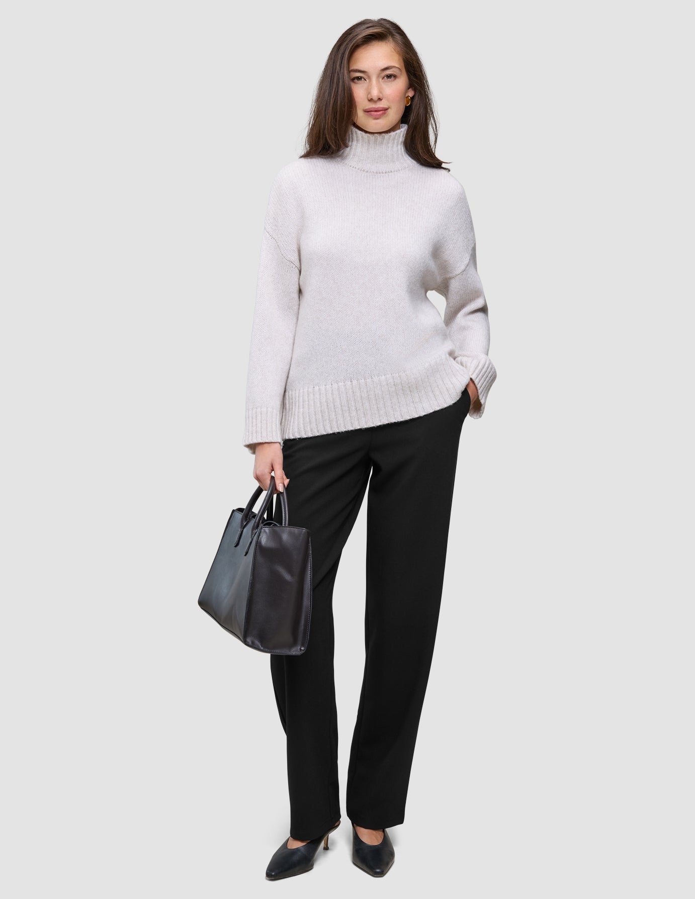 En kvinde i en beige rullekravesweater og et par sorte Women GO2-bukser med høj talje i lige snit, der holder en sort håndtaske.