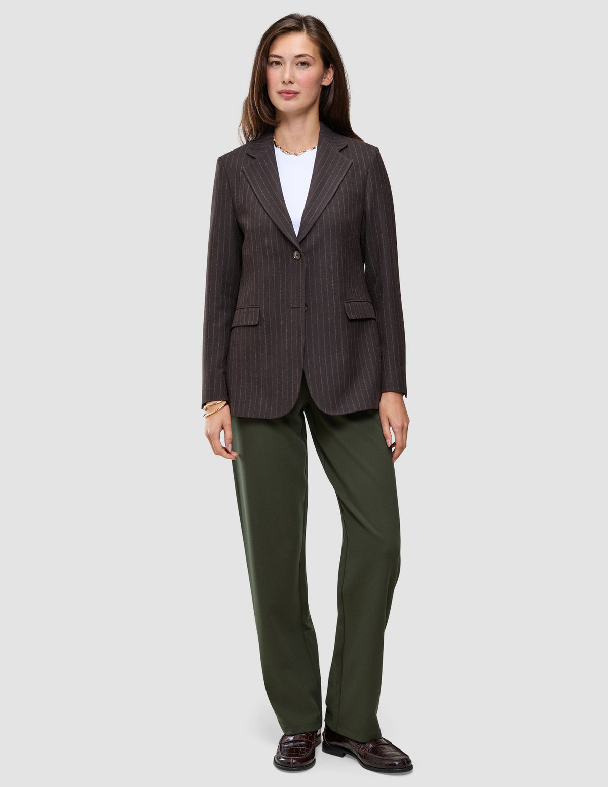 En kvinde har en brun blazer med tynde striber på over en hvid T-shirt og et par Women GO2-bukser i model Straight i farven Dusty Olive.