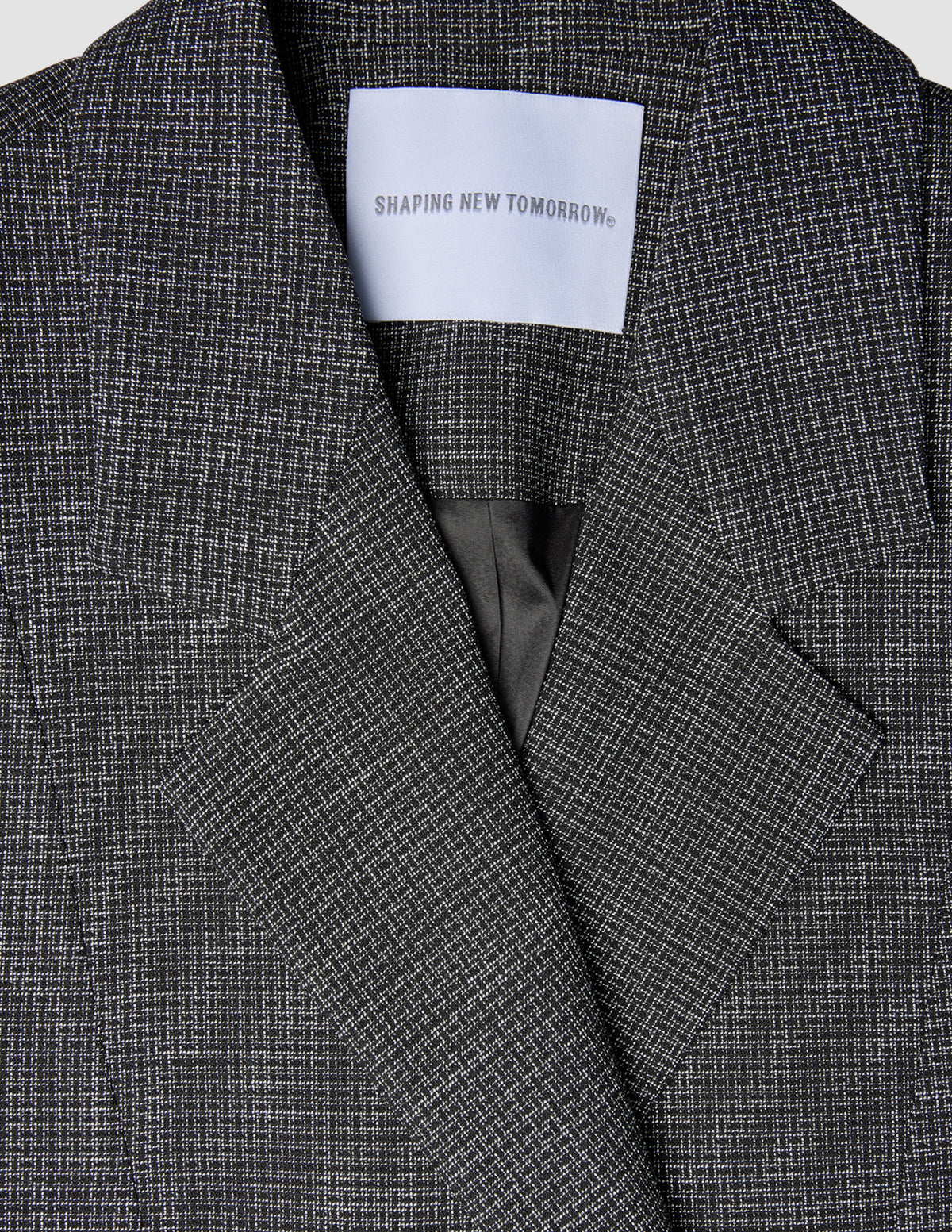 Nærbillede af en sort-hvid ternet Women Nara Blazer Fitted Black Check med et SHAPING NEW TOMORROW-mærke ved halsudskæringen.