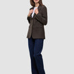 En kvinde har en brun Women Serene Blazer Regular Walnut Herringbone på over en hvid T-shirt, blå jeans og brune støvler.