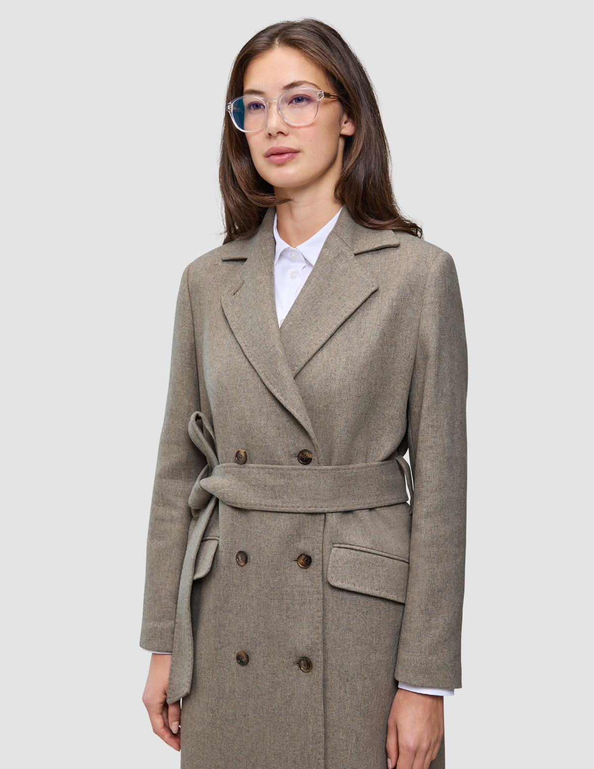 En kvinde har en Women Wool Coat Greige på – en lys brun-grå uldfrakke med dobbeltradet knapning – over en hvid skjorte med krave.