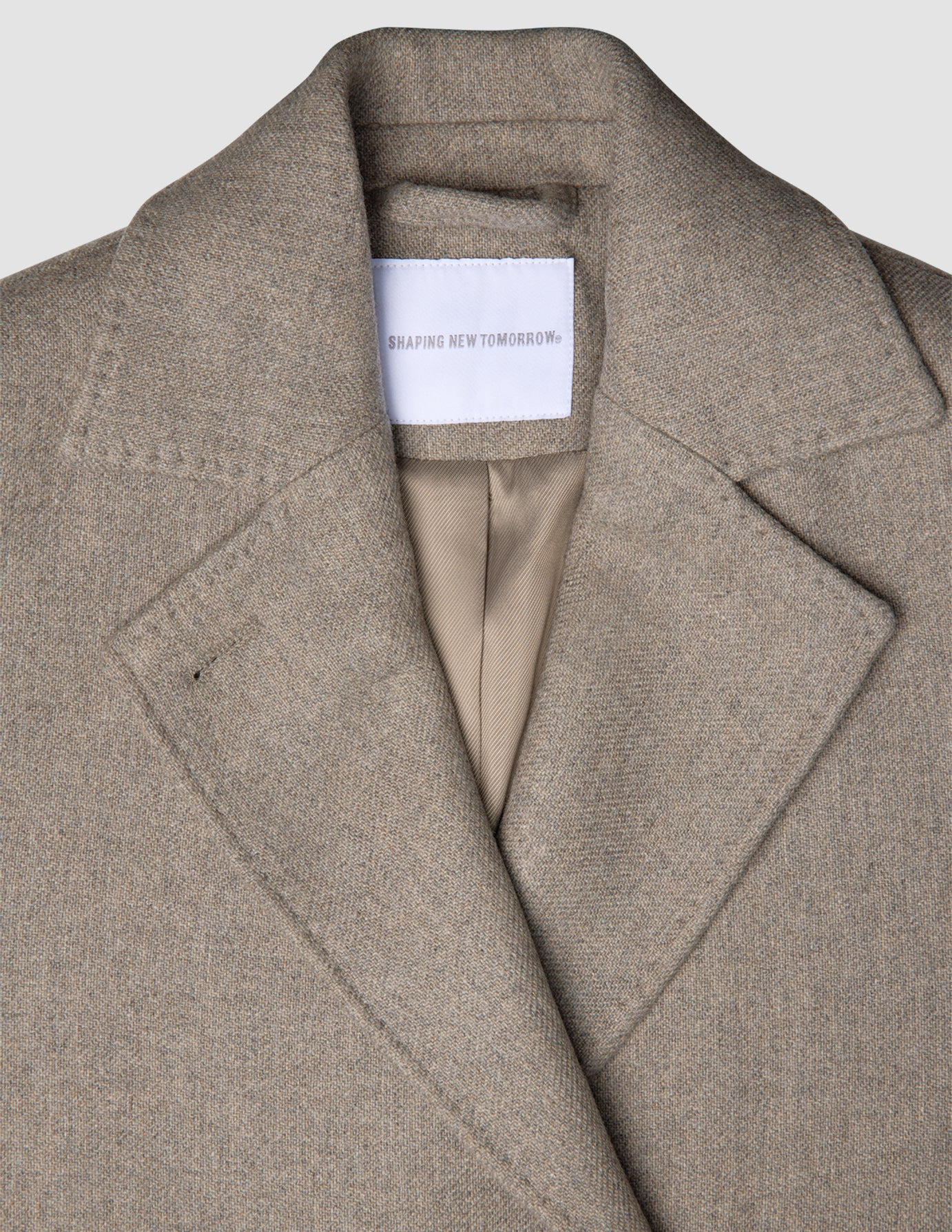 Nærbillede af en gråbeige uldfrakke til kvinder med skrå revers og et mærke på indermærket, der fremhæver klassisk dansk design.