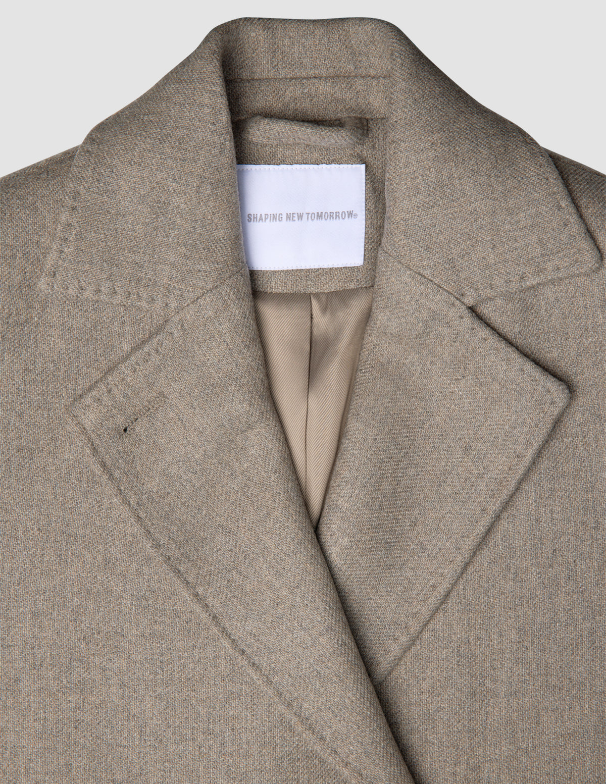 Nærbillede af en gråbeige uldfrakke til kvinder med skrå revers og et mærke på indermærket, der fremhæver klassisk dansk design.